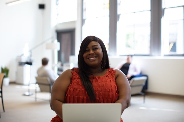 Woman in a red-orange dress using a laptop warmly smiles at the camera.