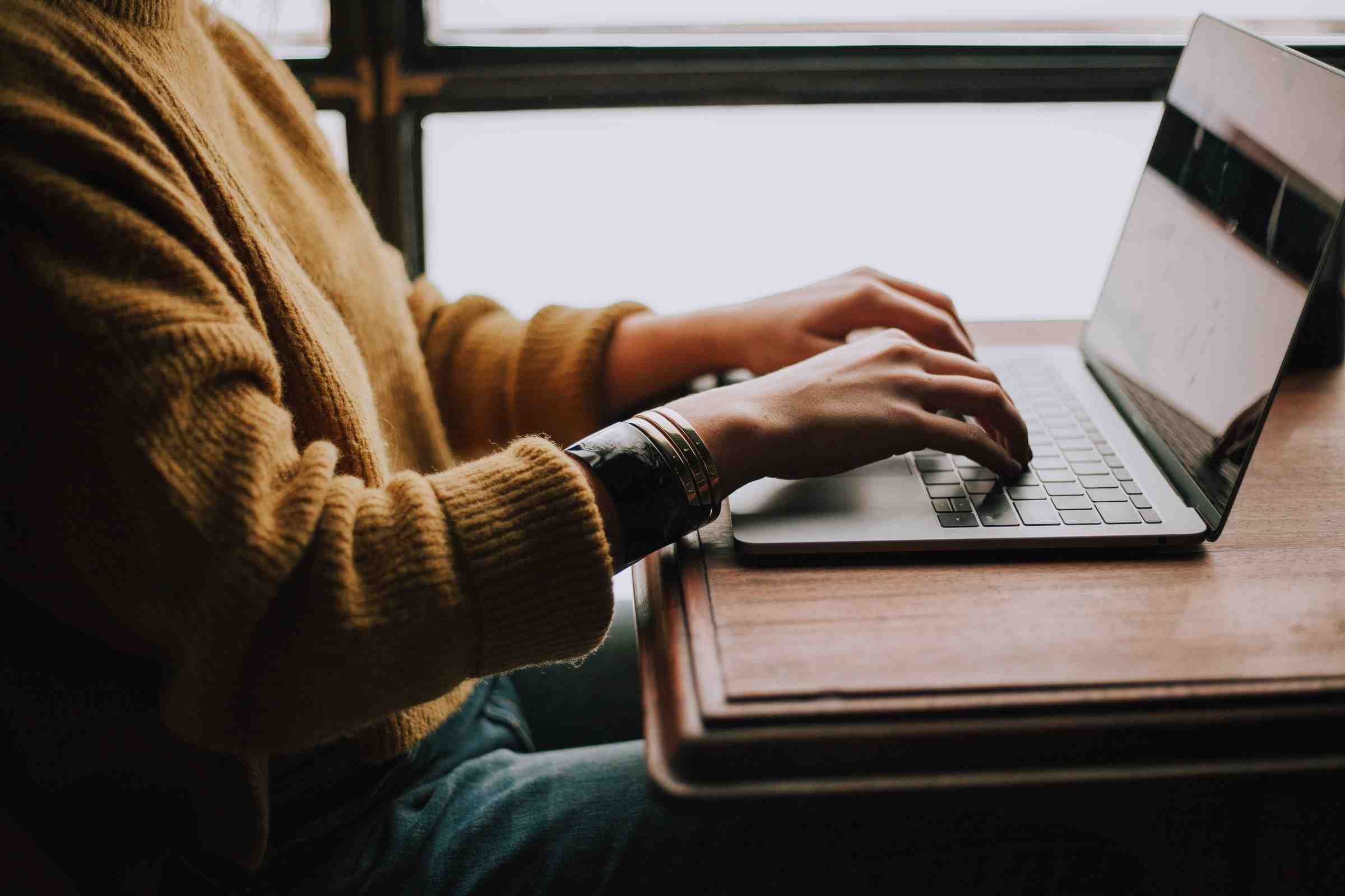 Person in a yellow wool sweater sits at a desk with their hand on a laptop keyboard. 